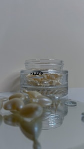 A clear jar containing pearl-like beauty capsules is labeled with 'KLAPP Beauty Capsules Protecting Serum + Vitamin E'. The jar is placed on a reflective surface, with several capsules scattered around, also reflected on the surface.