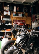 A cluttered garage workspace with two black motorcycles parked side by side. Shelves in the background are filled with various automotive fluids, spray cans, and maintenance tools. A Kawasaki sign is displayed on the wall, surrounded by a collection of posters and artwork. The scene is dimly lit, highlighting the metallic sheen of the motorcycles.