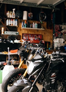 A cluttered garage workspace with two black motorcycles parked side by side. Shelves in the background are filled with various automotive fluids, spray cans, and maintenance tools. A Kawasaki sign is displayed on the wall, surrounded by a collection of posters and artwork. The scene is dimly lit, highlighting the metallic sheen of the motorcycles.