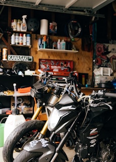 A cluttered garage workspace with two black motorcycles parked side by side. Shelves in the background are filled with various automotive fluids, spray cans, and maintenance tools. A Kawasaki sign is displayed on the wall, surrounded by a collection of posters and artwork. The scene is dimly lit, highlighting the metallic sheen of the motorcycles.