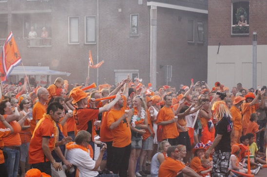 A large group of people gathered outside, wearing predominantly orange clothing and accessories such as hats and scarves. Many are holding flags and banners. The setting appears to be festive, with participants cheering and raising their hands. The background features residential buildings with some spectators watching from balconies.
