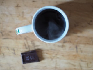 A close-up of a glossy chocolate bar next to a steaming cup of coffee, both styled to look delicious and inviting.