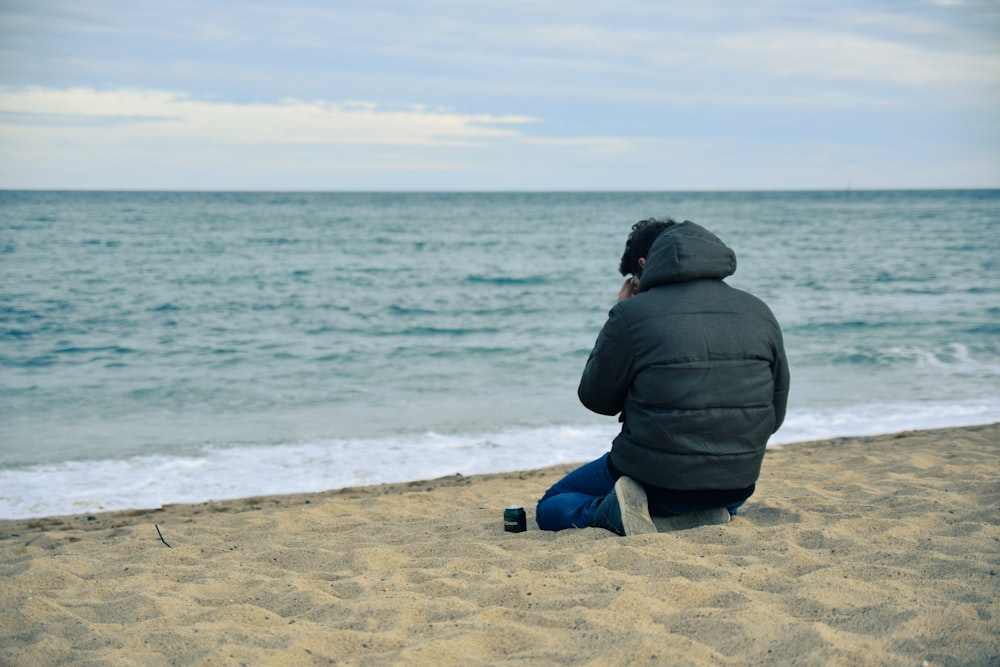homme en sweat à capuche noir et jean bleu assis sur le sable brun près d’un plan d’eau