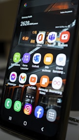 A close-up view of a smartphone screen displaying various application icons including Office, Teams, Camera, Messenger, Gmail, YouTube, and more. The background contains a reflection of an interior space with lighting and signage. There are also notifications and health data displayed at the top of the screen.