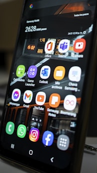 A close-up view of a smartphone screen displaying various application icons including Office, Teams, Camera, Messenger, Gmail, YouTube, and more. The background contains a reflection of an interior space with lighting and signage. There are also notifications and health data displayed at the top of the screen.