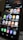 A close-up view of a smartphone screen displaying various application icons including Office, Teams, Camera, Messenger, Gmail, YouTube, and more. The background contains a reflection of an interior space with lighting and signage. There are also notifications and health data displayed at the top of the screen.