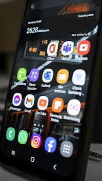 A close-up view of a smartphone screen displaying various application icons including Office, Teams, Camera, Messenger, Gmail, YouTube, and more. The background contains a reflection of an interior space with lighting and signage. There are also notifications and health data displayed at the top of the screen.