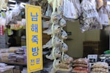 A market stall displays a variety of dried fish hanging vertically in bunches. Bags of dried goods are visible in the background, and a yellow sign with blue Korean text stands prominently in the foreground.