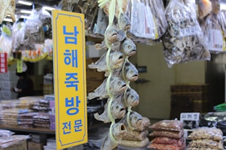 A market stall displays a variety of dried fish hanging vertically in bunches. Bags of dried goods are visible in the background, and a yellow sign with blue Korean text stands prominently in the foreground.