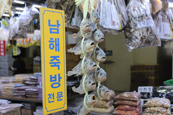 A market stall displays a variety of dried fish hanging vertically in bunches. Bags of dried goods are visible in the background, and a yellow sign with blue Korean text stands prominently in the foreground.