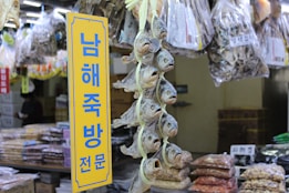 A market stall displays a variety of dried fish hanging vertically in bunches. Bags of dried goods are visible in the background, and a yellow sign with blue Korean text stands prominently in the foreground.