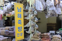 A market stall displays a variety of dried fish hanging vertically in bunches. Bags of dried goods are visible in the background, and a yellow sign with blue Korean text stands prominently in the foreground.