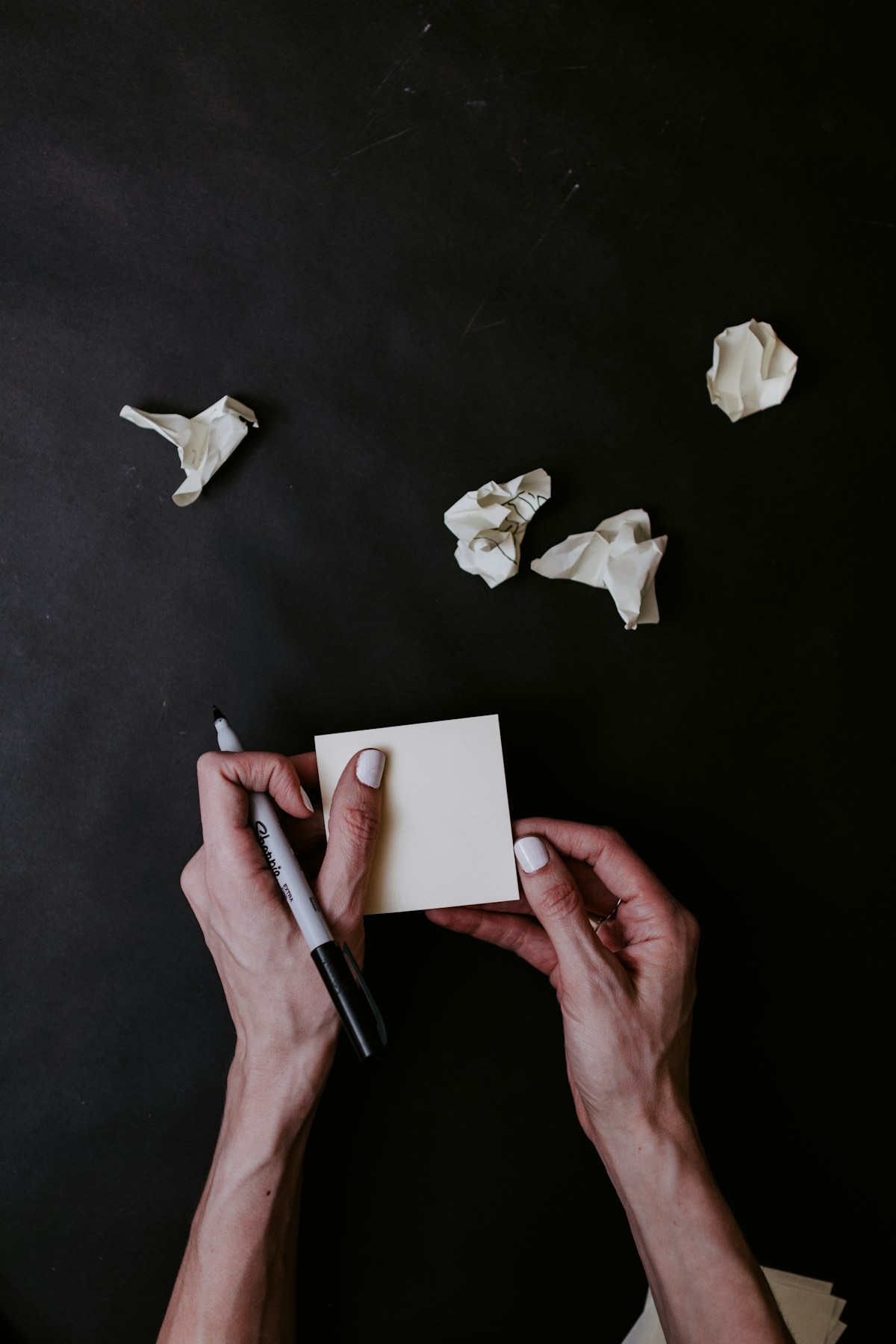 Two hands with white nail polish holding a pad of sticky notes and a sharpie. against a black background.  There are four crumpled notes lying in front of the hands.