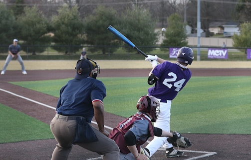 A baseball player in a purple jersey with the number 24 swings a bat, with an umpire and catcher in position nearby. The scene takes place on a baseball field with evergreen trees and a house in the background. The players and officials are focused on the game, indicating active play.