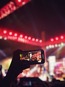 A person is recording a vibrant live music performance with a smartphone. The stage is illuminated with bright red and pink lights, and musicians can be seen performing in the background. The phone screen clearly displays the ongoing event, highlighting the intricate details of the stage setup, including instruments and sound equipment.