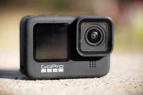 A compact action camera featuring a wide lens and a small display screen is positioned on a flat surface. The camera has a sleek black design with the brand name visible near the bottom.