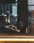 A bicycle is parked upside down indoors with its wheels elevated in the air. The setting appears to be a modern, dimly lit room with a large glass window on the right. The focus is on the bicycle, which has a sleek white frame and black wheels. The background features industrial architecture with muted colors.