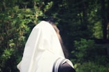 A person wearing a white prayer shawl is seen from the back, standing in a lush, green forest setting. The shawl has black stripes and covers most of the person's head and shoulders.