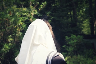 A person wearing a white prayer shawl is seen from the back, standing in a lush, green forest setting. The shawl has black stripes and covers most of the person's head and shoulders.