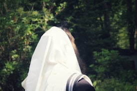 A person wearing a white prayer shawl is seen from the back, standing in a lush, green forest setting. The shawl has black stripes and covers most of the person's head and shoulders.