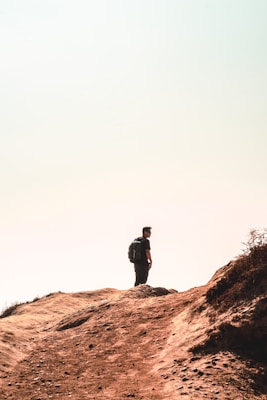 A lone person stands on a rocky, dusty trail with a backpack, gazing into the distance. The landscape is arid, with sparse vegetation and soft sunlight creating a hazy atmosphere.