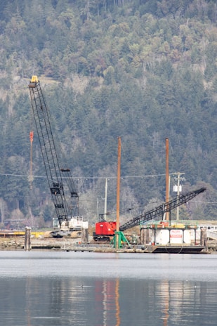 A construction site by the water with two large cranes, where various machinery and equipment are visible. The background features a dense, forested hillside with numerous trees.