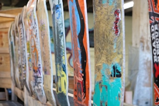 A row of well-used and colorful skateboard decks are lined up vertically, each displaying a variety of graphics and wear marks. The decks show signs of heavy use, with chipped paint and scratches. The background includes a wooden structure and some blurred elements, adding to the industrial and artistic feel.