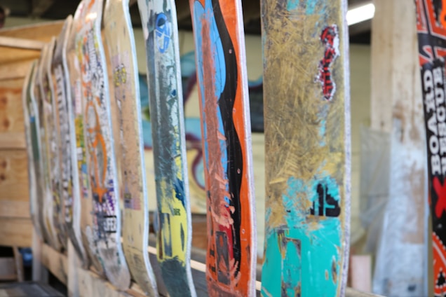 A row of well-used and colorful skateboard decks are lined up vertically, each displaying a variety of graphics and wear marks. The decks show signs of heavy use, with chipped paint and scratches. The background includes a wooden structure and some blurred elements, adding to the industrial and artistic feel.