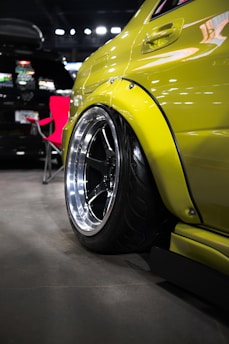 A close-up of the rear wheel of a customized yellow car with a glossy finish. The car is parked indoors beside a red folding chair and another vehicle, which is black. The wheel features a shiny rim and low-profile tire, emphasizing the car's modified appearance.