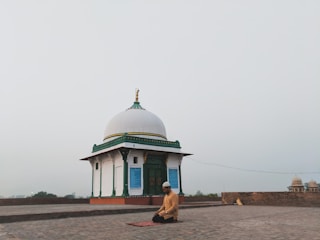 A person is kneeling in prayer on a mat in front of a small white domed building with green and gold accents. The setting is open and expansive, possibly on a rooftop or an open courtyard, with a hazy sky in the background.