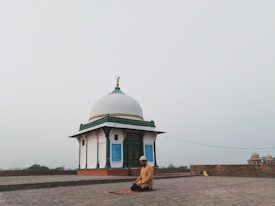 A person is kneeling in prayer on a mat in front of a small white domed building with green and gold accents. The setting is open and expansive, possibly on a rooftop or an open courtyard, with a hazy sky in the background.