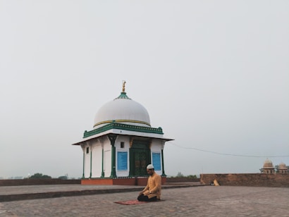 A person is kneeling in prayer on a mat in front of a small white domed building with green and gold accents. The setting is open and expansive, possibly on a rooftop or an open courtyard, with a hazy sky in the background.