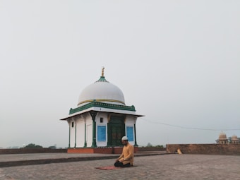 A person is kneeling in prayer on a mat in front of a small white domed building with green and gold accents. The setting is open and expansive, possibly on a rooftop or an open courtyard, with a hazy sky in the background.