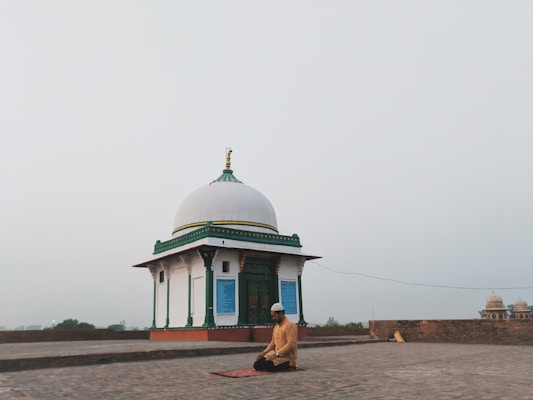 A person is kneeling in prayer on a mat in front of a small white domed building with green and gold accents. The setting is open and expansive, possibly on a rooftop or an open courtyard, with a hazy sky in the background.