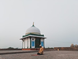 A person is kneeling in prayer on a mat in front of a small white domed building with green and gold accents. The setting is open and expansive, possibly on a rooftop or an open courtyard, with a hazy sky in the background.