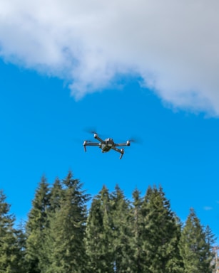 A drone hovers in the clear blue sky above a dense forest of tall evergreen trees. White clouds are scattered across the sky, and the drone is positioned in the center of the image.