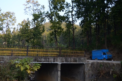 A blue delivery truck is parked on a dirt road near a concrete bridge. Tall trees with green foliage surround the area, casting shade and creating a serene, natural environment. The bridge has yellow railings, and lush vegetation is visible in the foreground.