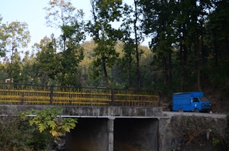 A blue delivery truck is parked on a dirt road near a concrete bridge. Tall trees with green foliage surround the area, casting shade and creating a serene, natural environment. The bridge has yellow railings, and lush vegetation is visible in the foreground.