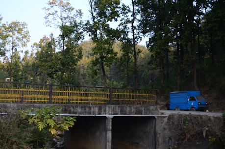 A blue delivery truck is parked on a dirt road near a concrete bridge. Tall trees with green foliage surround the area, casting shade and creating a serene, natural environment. The bridge has yellow railings, and lush vegetation is visible in the foreground.