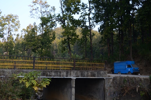 A blue delivery truck is parked on a dirt road near a concrete bridge. Tall trees with green foliage surround the area, casting shade and creating a serene, natural environment. The bridge has yellow railings, and lush vegetation is visible in the foreground.