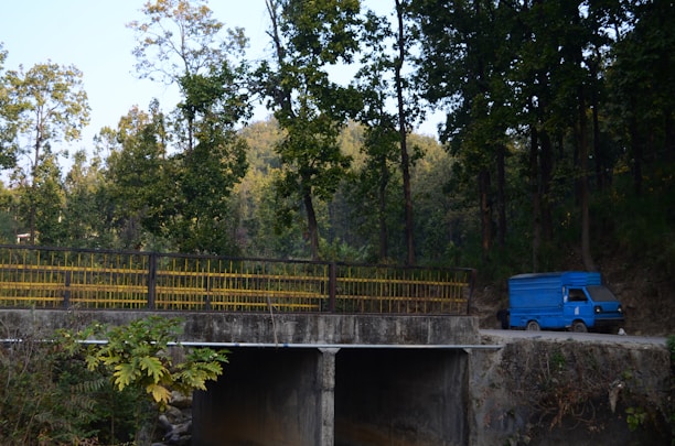 A blue delivery truck is parked on a dirt road near a concrete bridge. Tall trees with green foliage surround the area, casting shade and creating a serene, natural environment. The bridge has yellow railings, and lush vegetation is visible in the foreground.