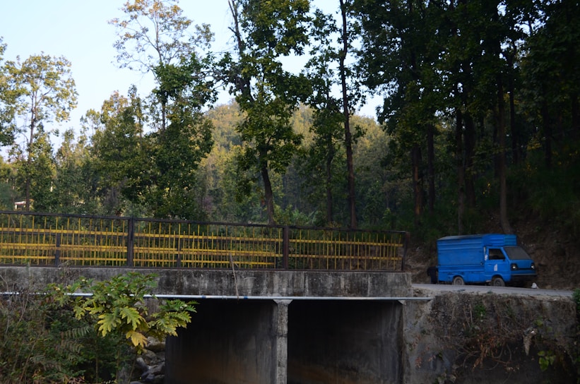A blue delivery truck is parked on a dirt road near a concrete bridge. Tall trees with green foliage surround the area, casting shade and creating a serene, natural environment. The bridge has yellow railings, and lush vegetation is visible in the foreground.