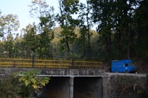 A blue delivery truck is parked on a dirt road near a concrete bridge. Tall trees with green foliage surround the area, casting shade and creating a serene, natural environment. The bridge has yellow railings, and lush vegetation is visible in the foreground.