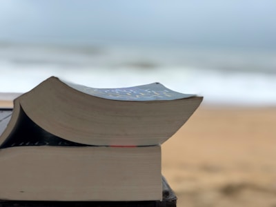A close-up view of two stacked books with slightly curved pages, set against a blurred beach and ocean background.