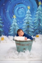 A sleeping baby dressed in blue is nestled in a rustic metal bucket, surrounded by fluffy white material resembling snow. The background features a whimsical painted scene with swirling dark blue skies and evergreen trees topped with stars, creating a serene, wintery atmosphere.