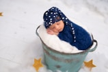 A newborn baby is peacefully sleeping in a blue, fuzzy wrap and a matching hat with white stars, placed inside a rustic, teal bucket lined with soft white material. The setting includes a fluffy white surface with yellow star decorations scattered around, creating a dreamy and serene atmosphere.