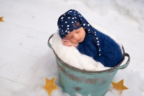 A newborn baby is peacefully sleeping in a blue, fuzzy wrap and a matching hat with white stars, placed inside a rustic, teal bucket lined with soft white material. The setting includes a fluffy white surface with yellow star decorations scattered around, creating a dreamy and serene atmosphere.