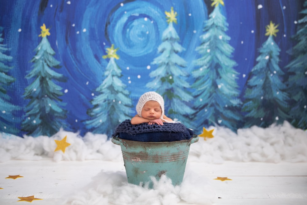 child in black jacket and white knit cap sitting on blue plastic bucket on snow covered