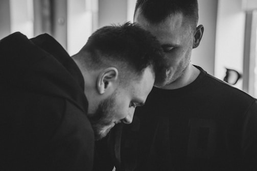 Two men are close together, partially embracing or leaning towards each other, with soft lighting and a blurred background adding a sense of intimacy.