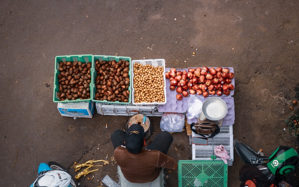 man in black jacket sitting on the floor with red round fruits on the crate